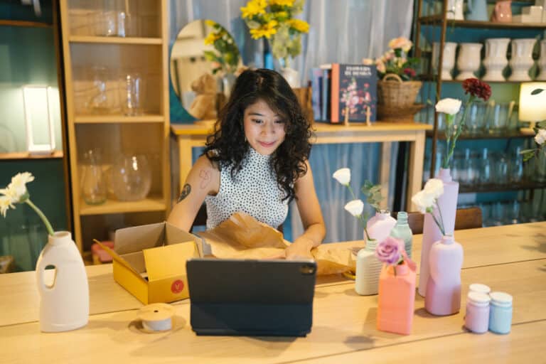 Businesswoman using laptop for ecommerce product discovery while packing vase from plastic in a cardboard box.