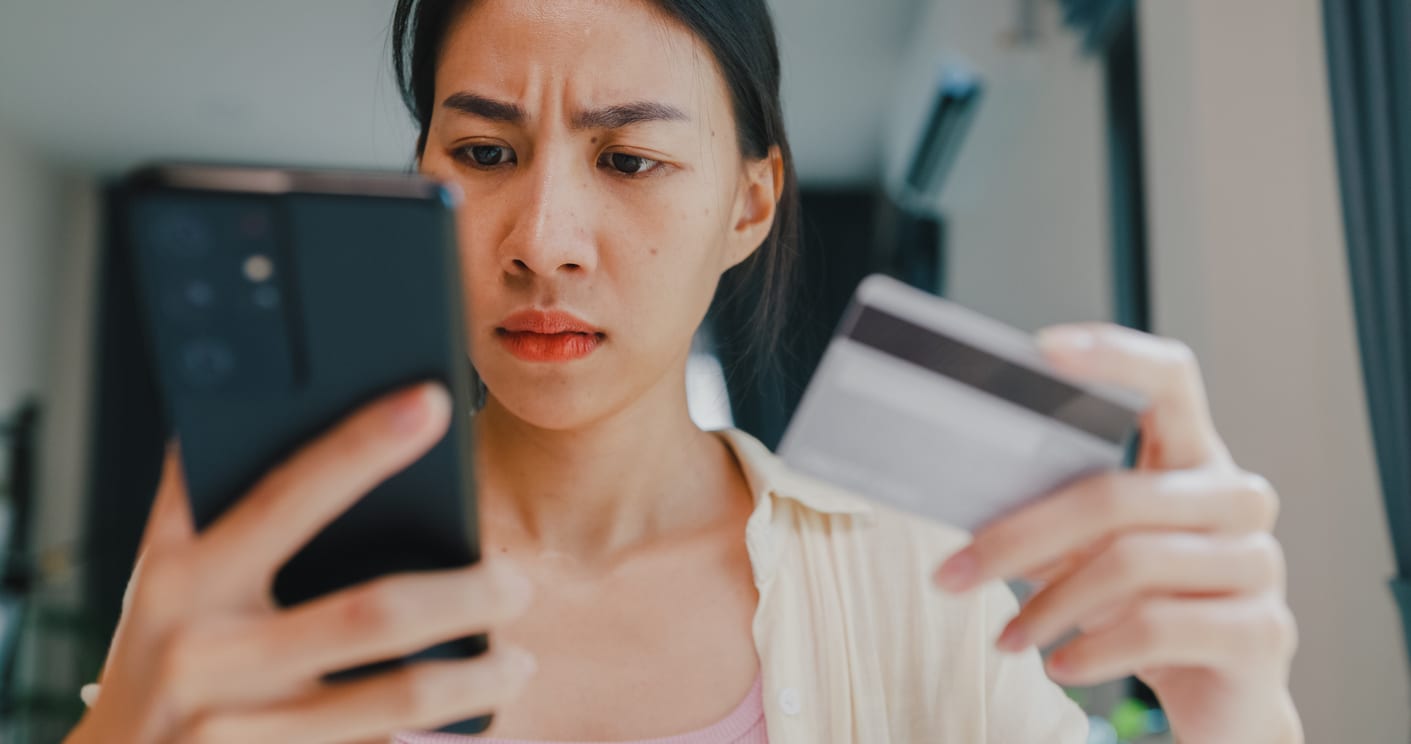 A frustrated woman looks at her phone and credit card, confused by fluctuating prices or complex online shopping procedures, highlighting the potential challenges of dynamic pricing for consumers.