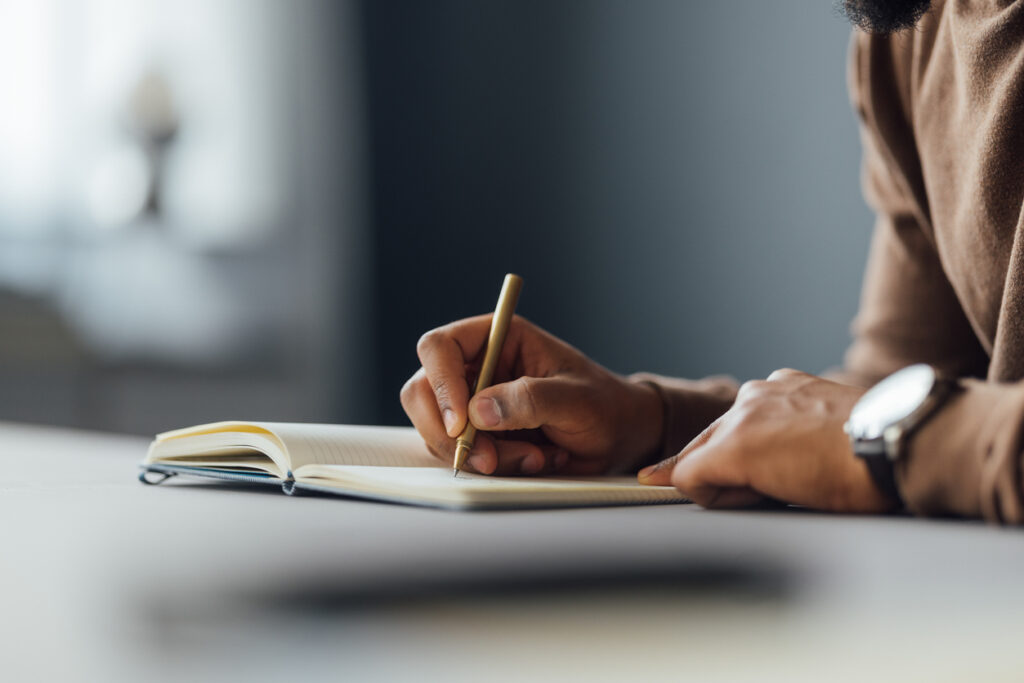 A close-up image of a man writing in a notebook with a pen at a desk, focusing on creativity and concentration.