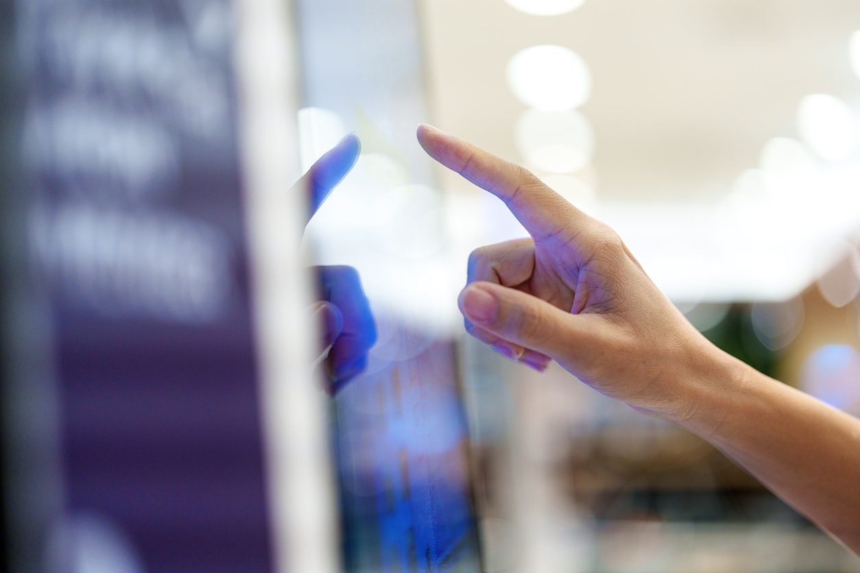 A woman is seen from the side, using a touch screen display to confirm an order or payment at a kiosk machine in a shopping mall. This concept highlights the future of service delivery, emphasizing the role of kiosks in improving the customer experience