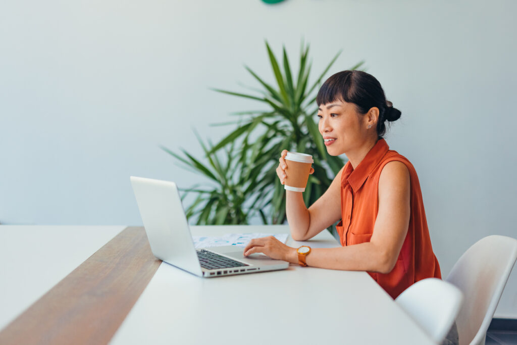 A professional Asian woman in an orange blouse is engaged in an online meeting, smiling while holding a coffee cup. She is sitting at a desk with a laptop and paperwork.