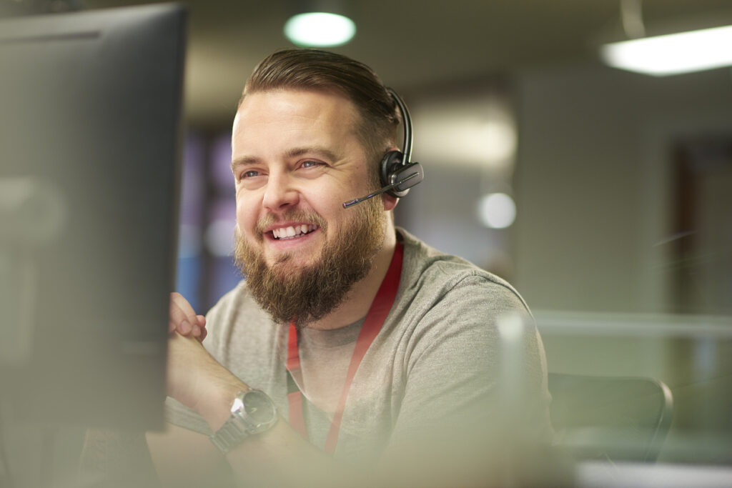 a call centre phone operative in his mid 30s chats on the phone at his desk . He is explaining something to the person on the phone in a friendly manner . behind him a defocussed office interior can be seen . This could be a call centre or an office worker chatting to a customer .