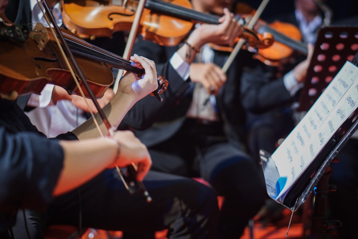 Orchestra musicians playing violins with sheet music, illustrating how AI merchandising platforms require human expertise and interpretation similar to how musicians execute a musical score under a conductor's guidance.